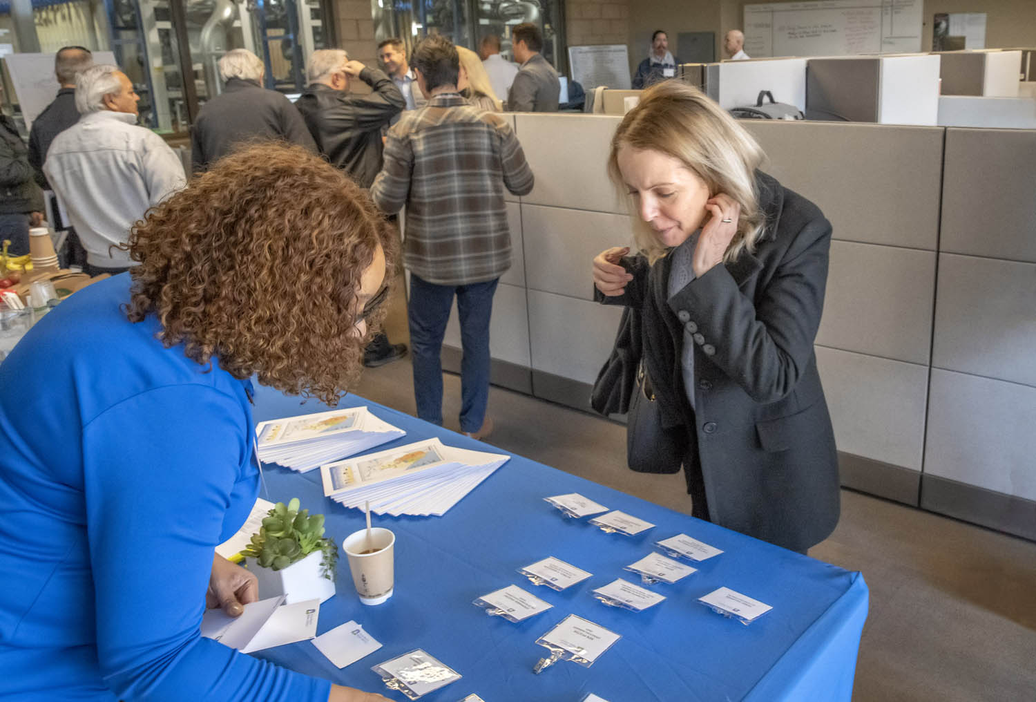 Two women at a registration table with badges, brochures, and a small plant in a busy office lobby.