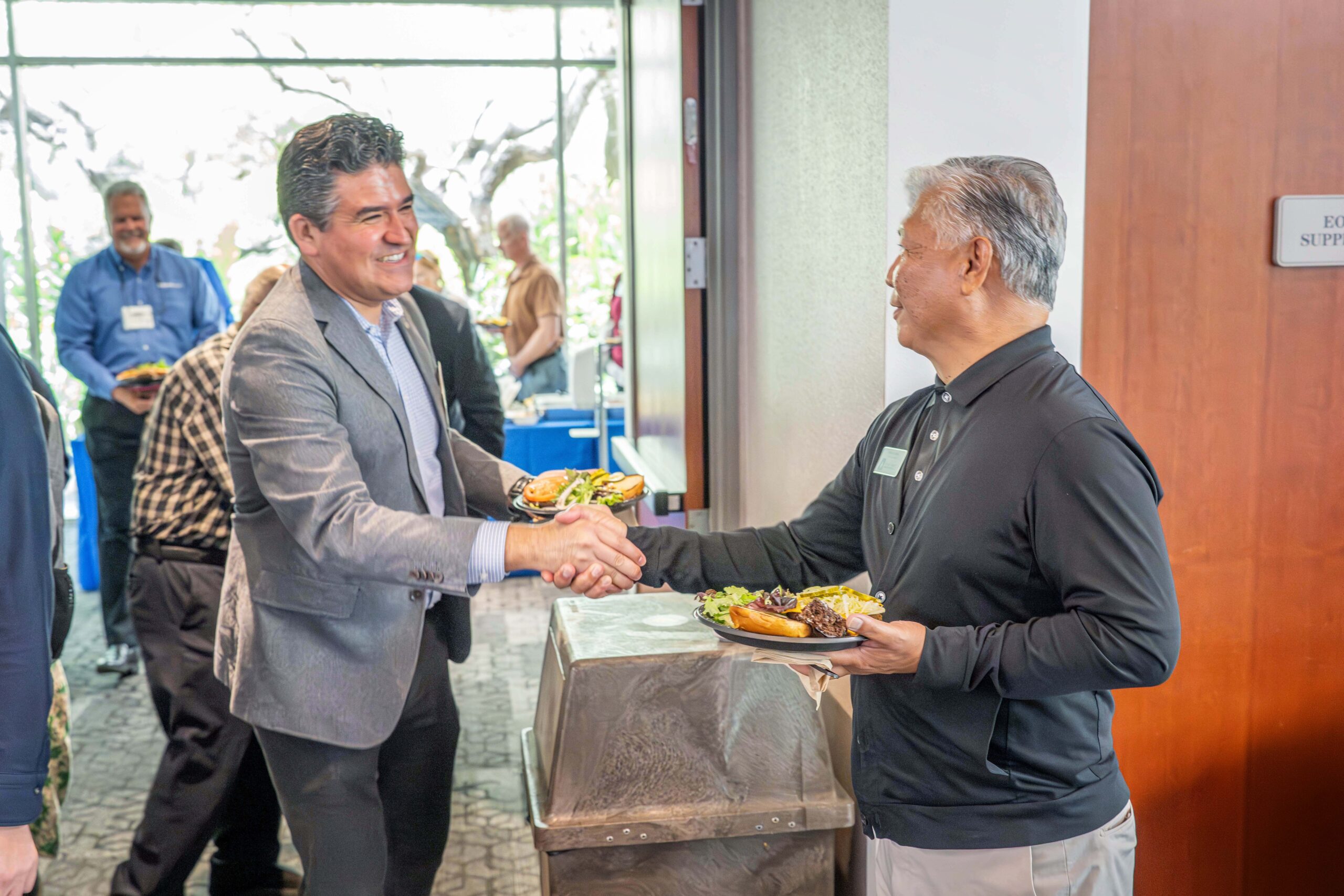 Two men shake hands at a buffet during a social event; one wears a gray blazer, the other a black polo.