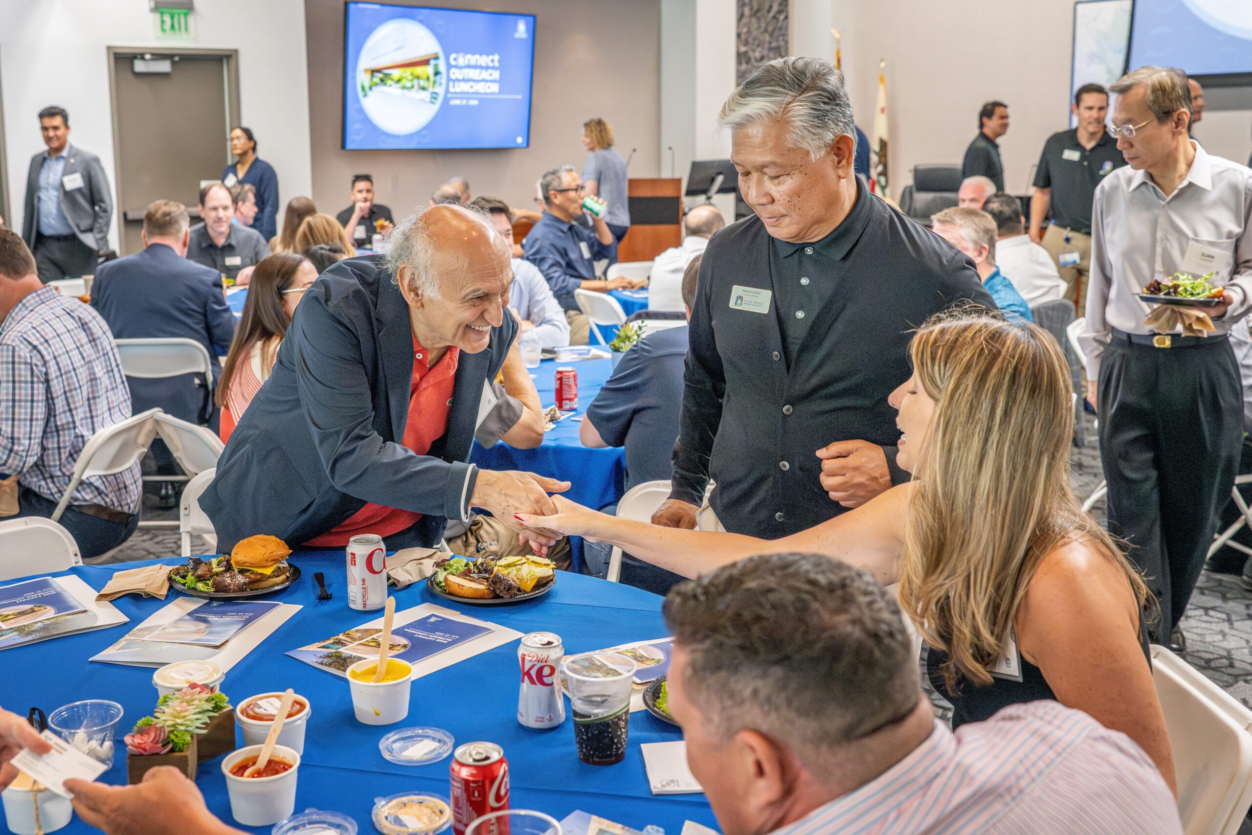 Two attendees shake hands across a round table at a crowded conference luncheon.