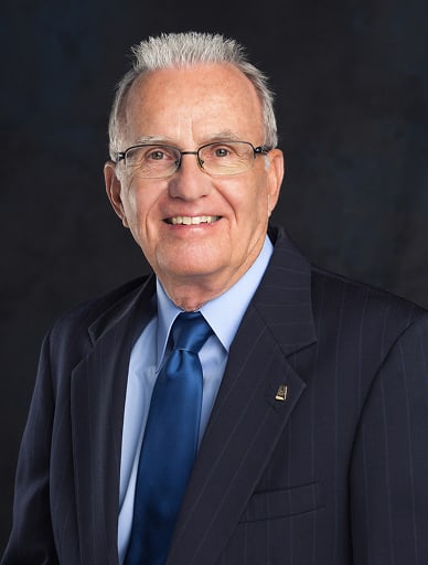 Senior man in a dark pinstripe suit, blue tie, and glasses, smiling.