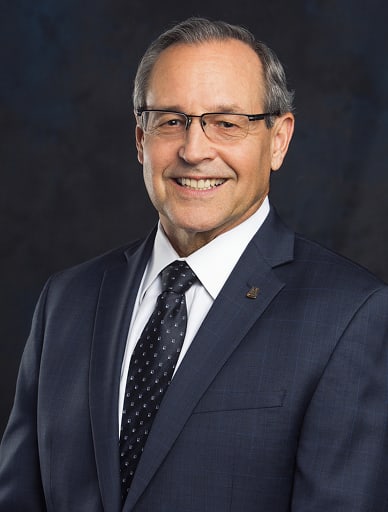 Smiling middle-aged man in a navy suit and dotted tie, wearing glasses, against a dark studio backdrop.