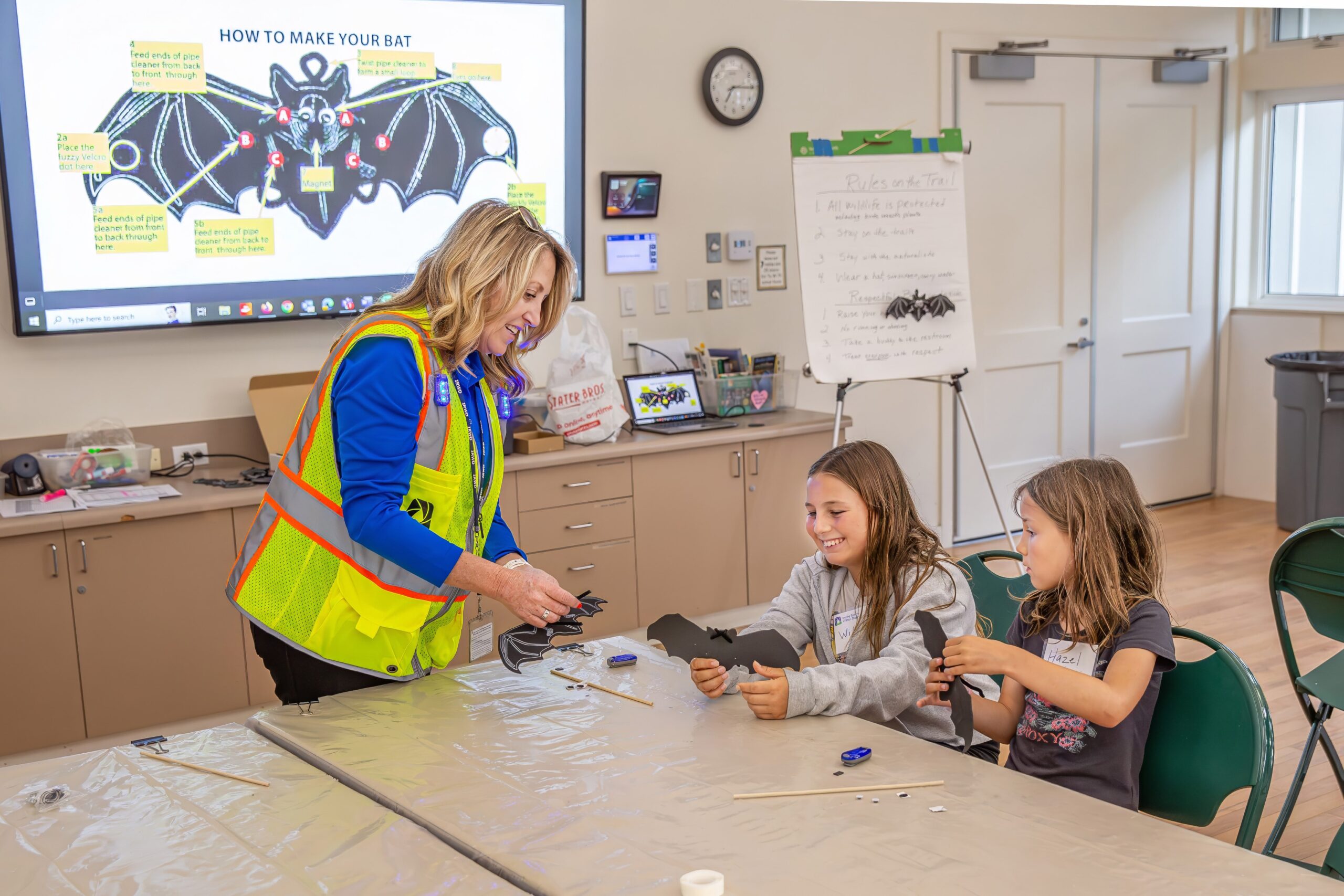 Instructor in a neon safety vest helps two kids cut out bat shapes at a classroom craft table.