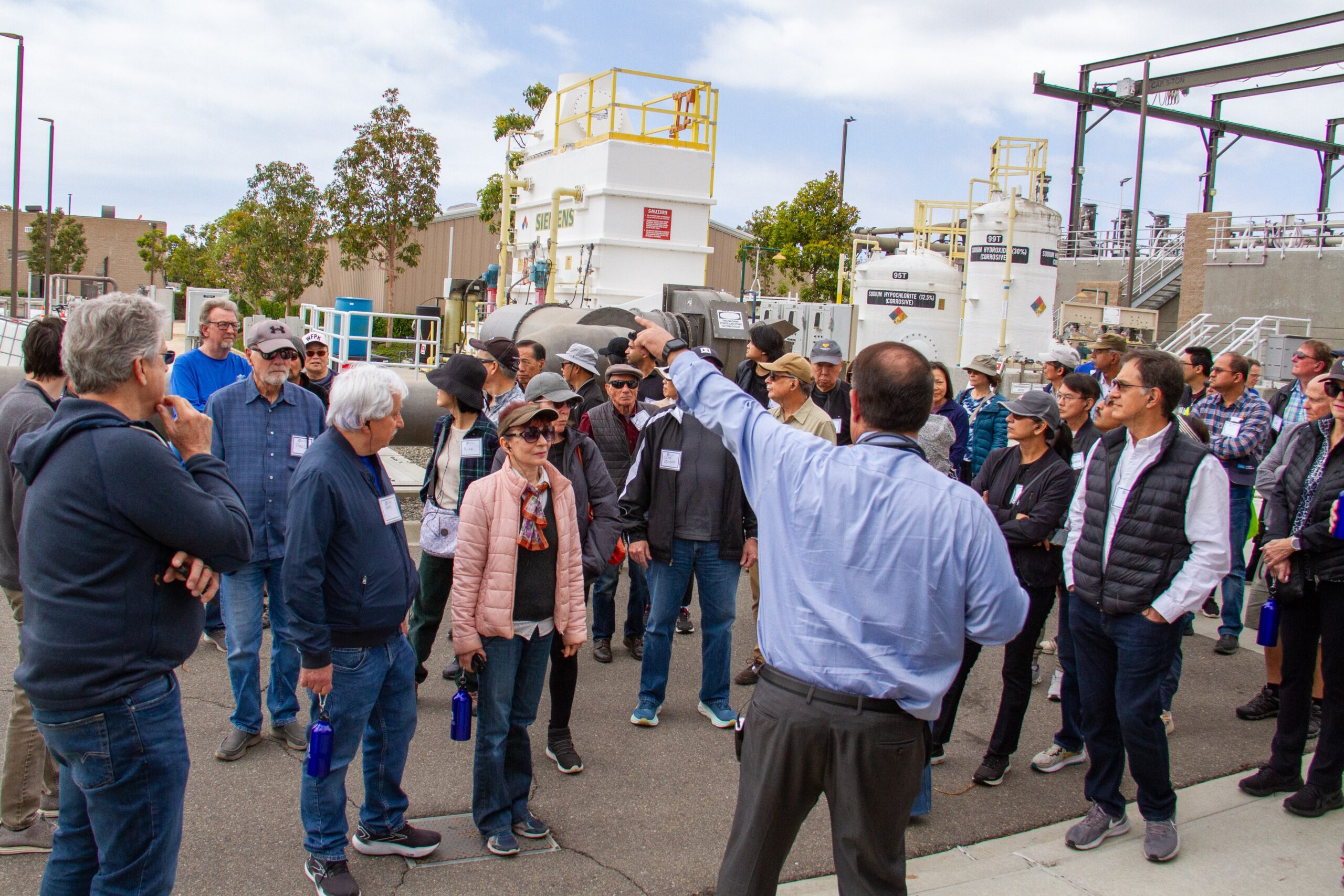 Group of adults on an industrial facility tour listening to a guide who points toward equipment.
