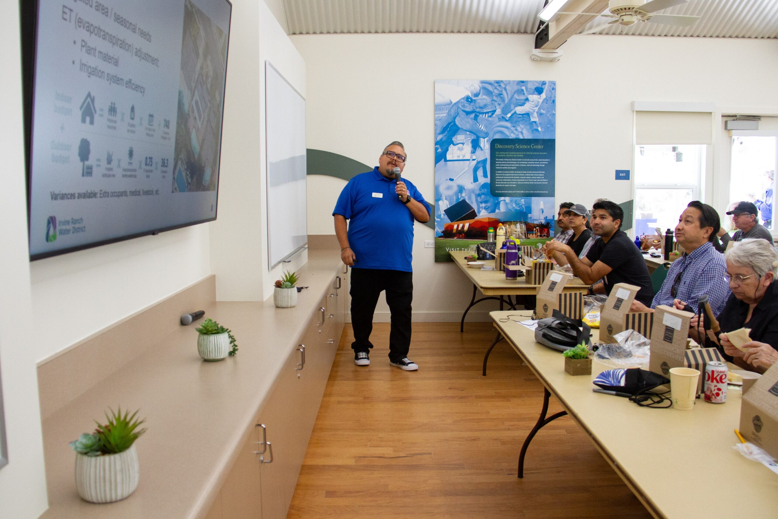 Presenter in a blue polo speaks into a microphone to a group seated at long tables in a classroom setting with a large screen.