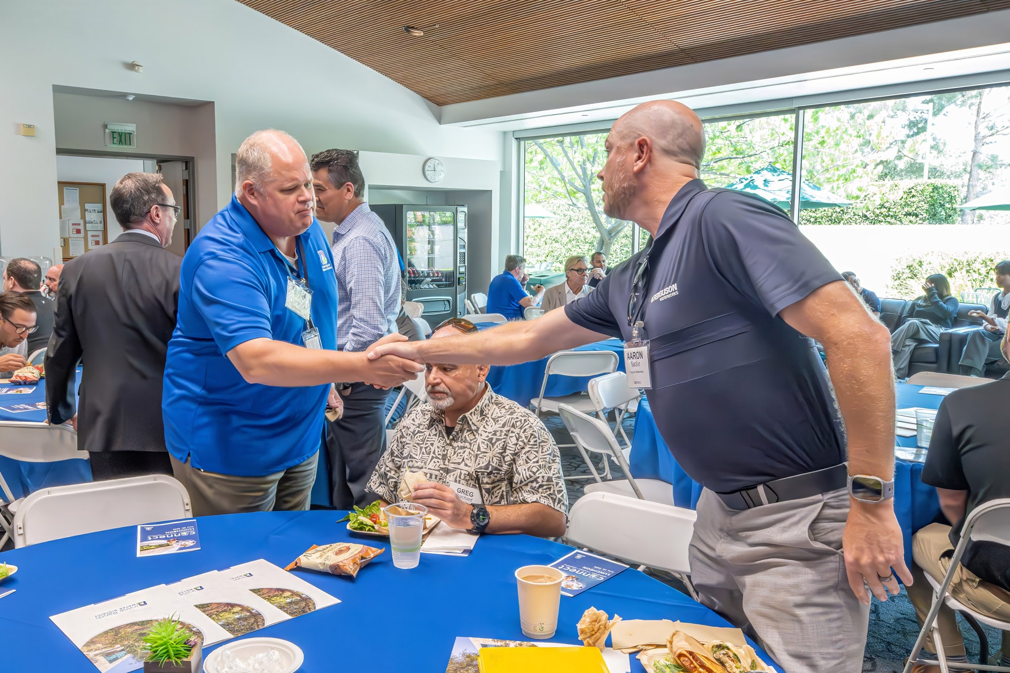 Two men shake hands across a table at a business networking event, with a seated participant between them.