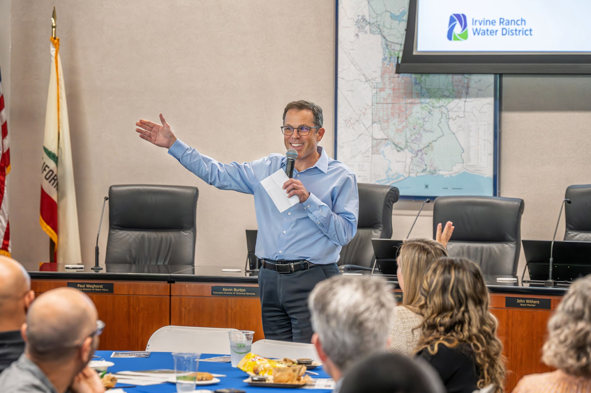 Man in a light blue shirt speaks at a meeting, holding a microphone, with a map and Irvine Ranch Water District logo visible behind.
