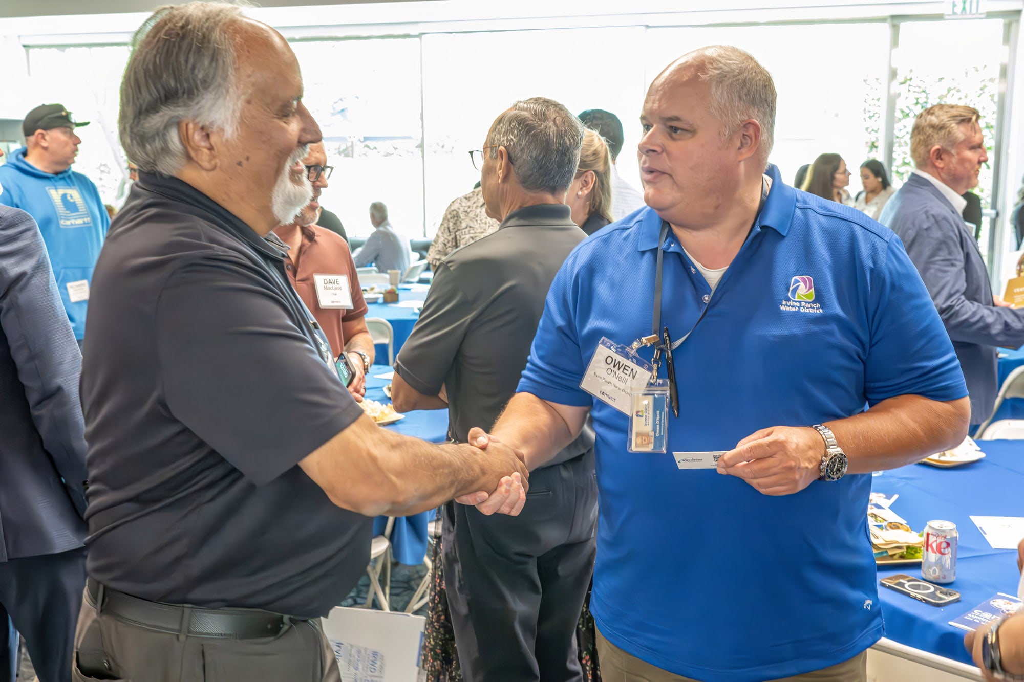 Two men shake hands at a networking event with blue tablecloths and attendees in the background.