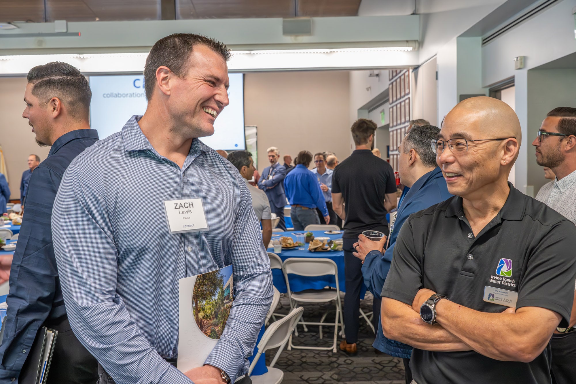 Smiling man in a light-blue checkered shirt talks with a colleague at a networking event; a name badge is visible.