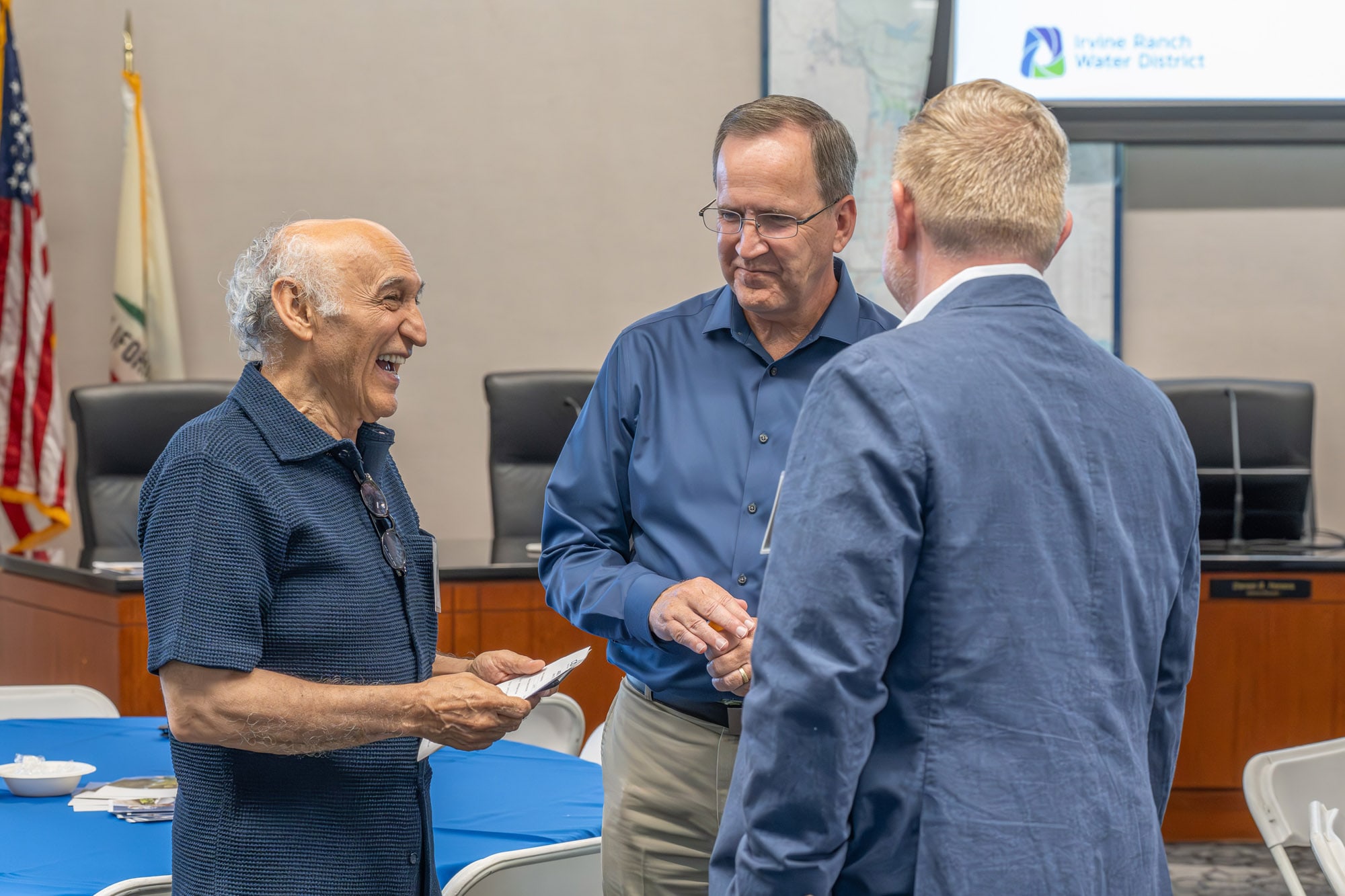Three men in a boardroom chat and laugh, with a blue table in front and a screen showing the Irvine Ranch Water District logo.