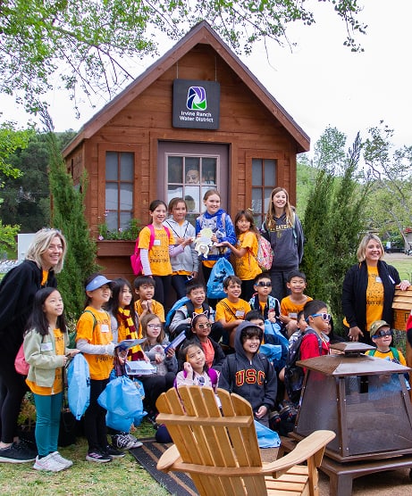 Group of diverse children in orange shirts posing in front of a wooden cabin with Irvine Ranch Water District sign during a field trip.