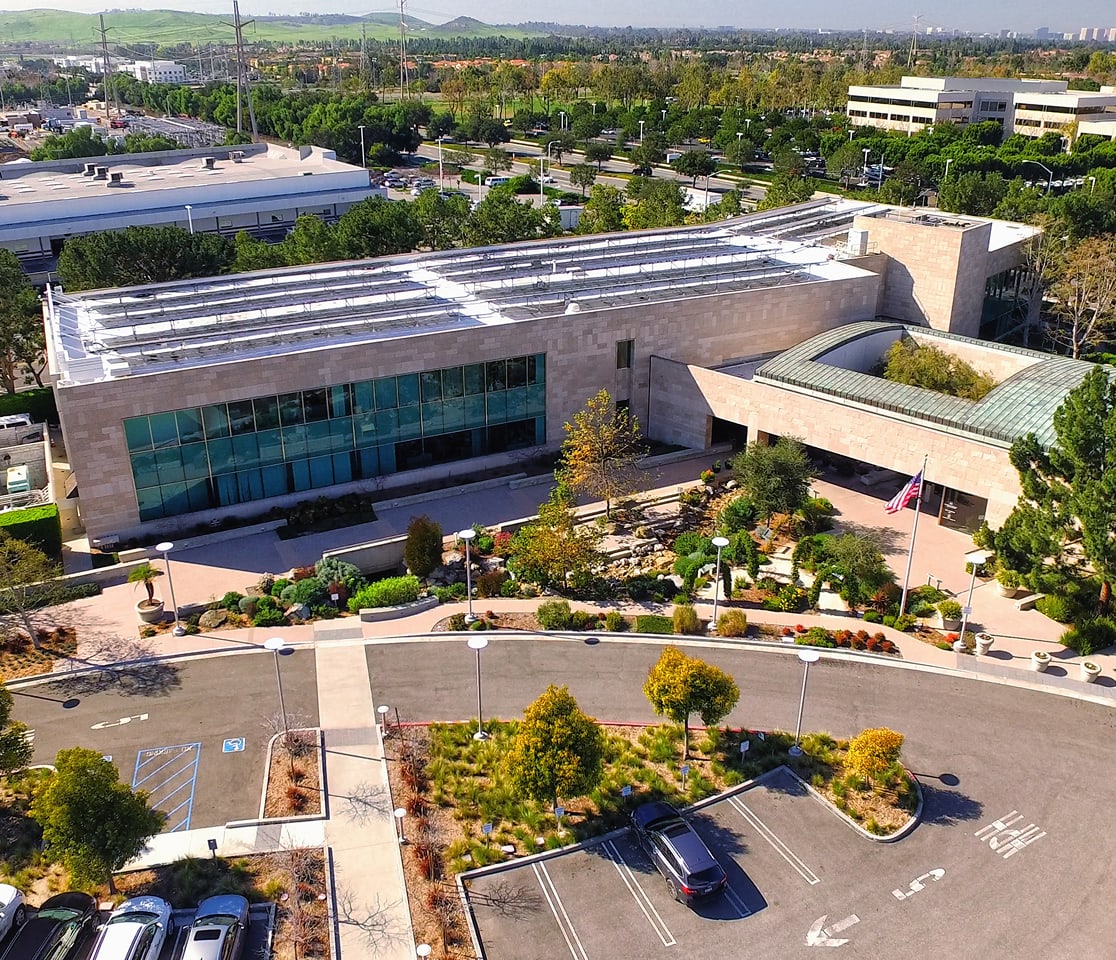 Aerial view of a modern campus building with glass facade, landscaped entrance, and parking lot.