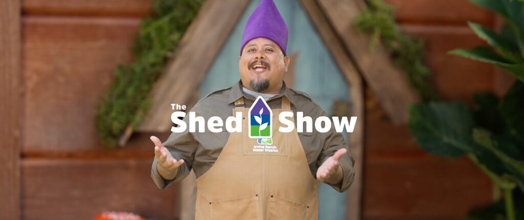 Man in a tan apron and purple cone-shaped hat gestures with open hands before a wooden shed backdrop.