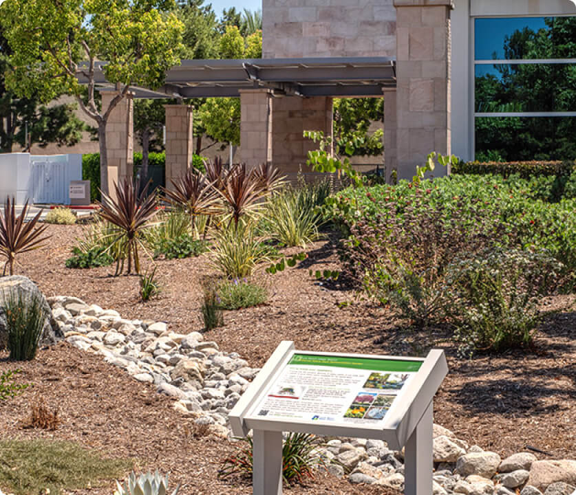 Desert-style landscape with drought-tolerant plants, rocks, and an informational sign by a modern building.
