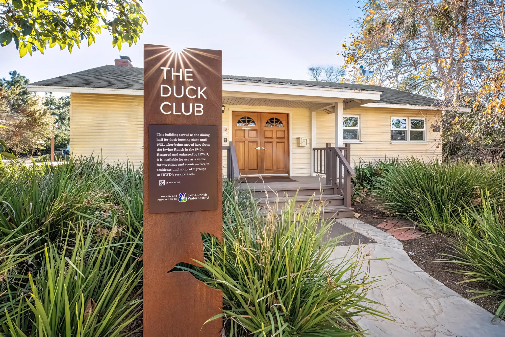 Front view of The Duck Club building with a tall rust-colored sign, wooden double doors, and lush landscaping