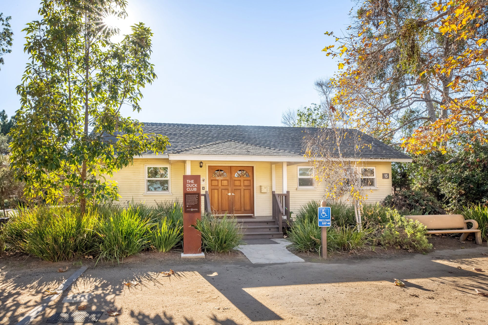 Small beige community building with wooden double doors labeled The Duck Club, surrounded by greenery and autumn trees.