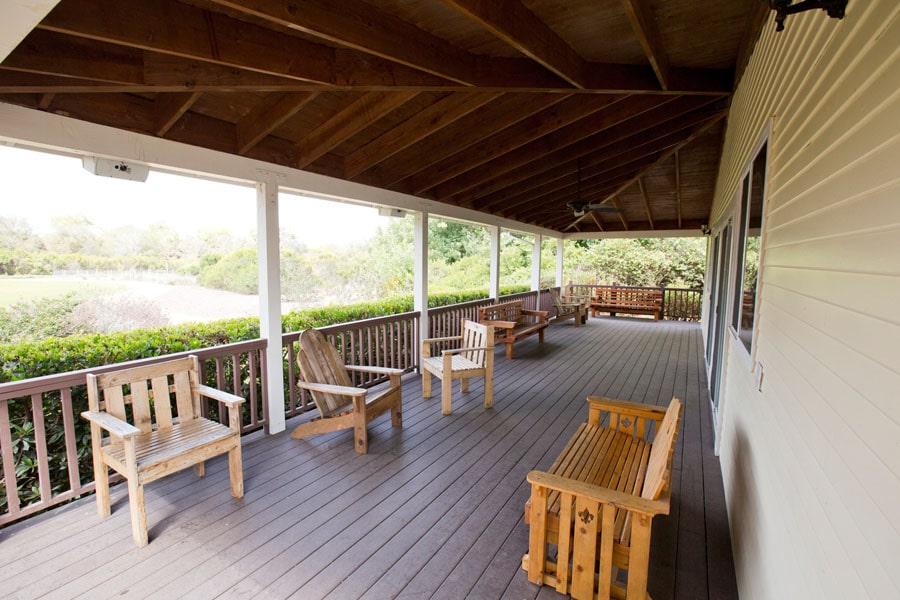 Covered wooden porch with multiple chairs and benches arranged along the railing overlooking a garden.