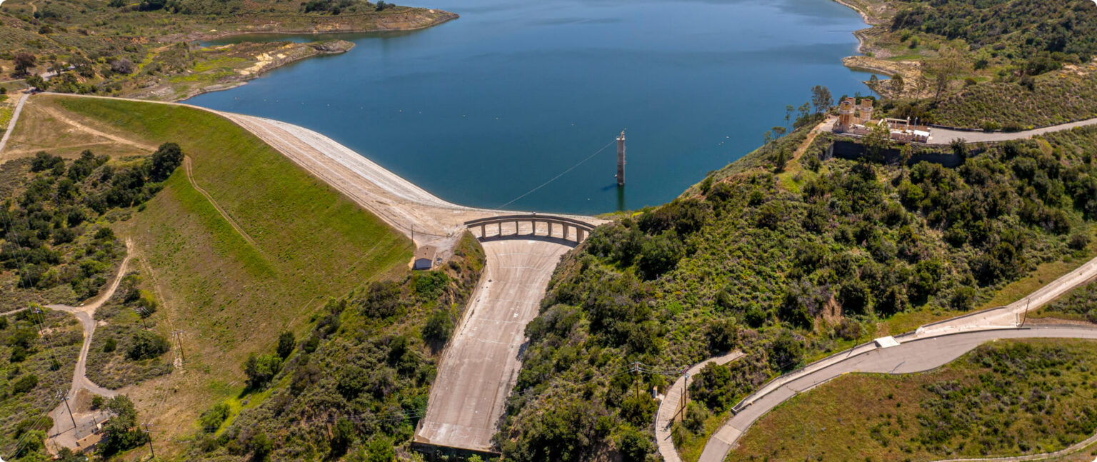 Aerial view of a dam across a curved valley with a wide spillway and blue reservoir, surrounded by green hills and roads.