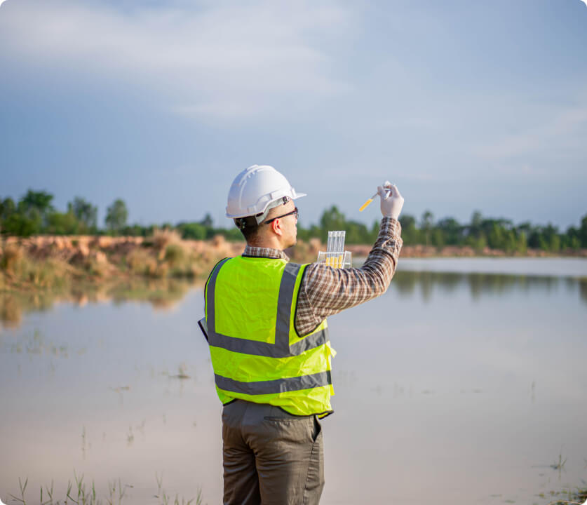 Construction worker in a neon safety vest and hard hat samples liquid with a dropper by a pond.