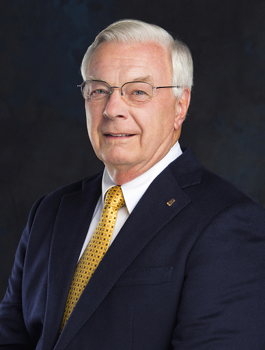 Professional portrait of an older man with silver hair and glasses wearing a navy suit and yellow tie.