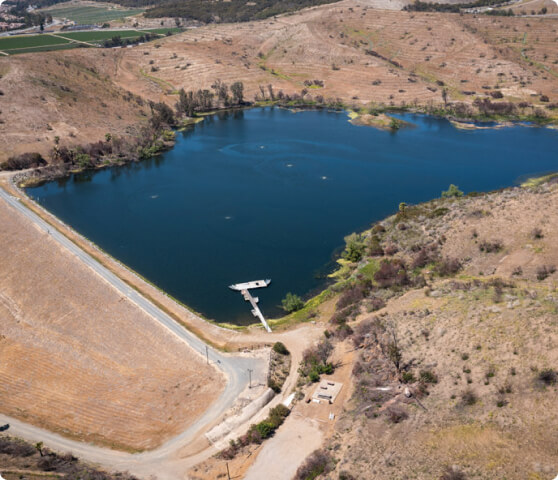 Aerial view of a deep blue reservoir nestled among dry brown hills, with a small dam and a road along the shore.