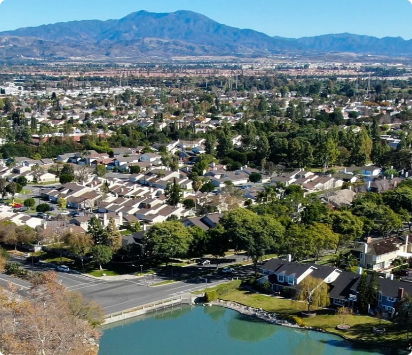 Aerial view of a suburban neighborhood with rows of houses, trees, a lake in the foreground, and mountains on the horizon.