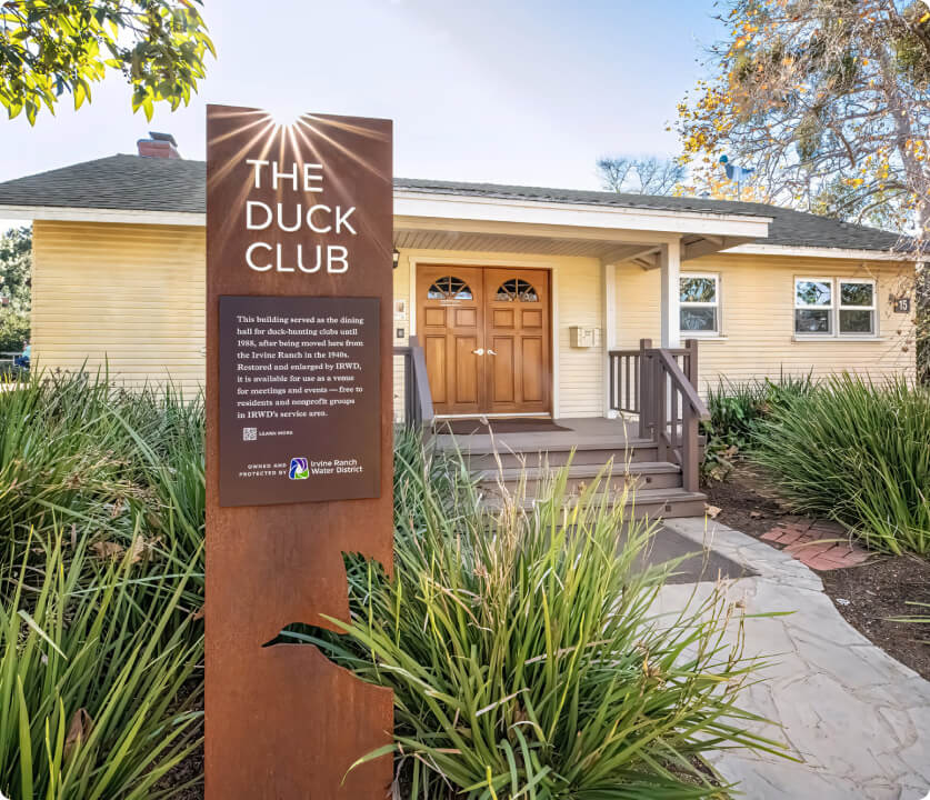 Rust-colored sign reading The Duck Club in front of a beige, single-story building with wooden doors.