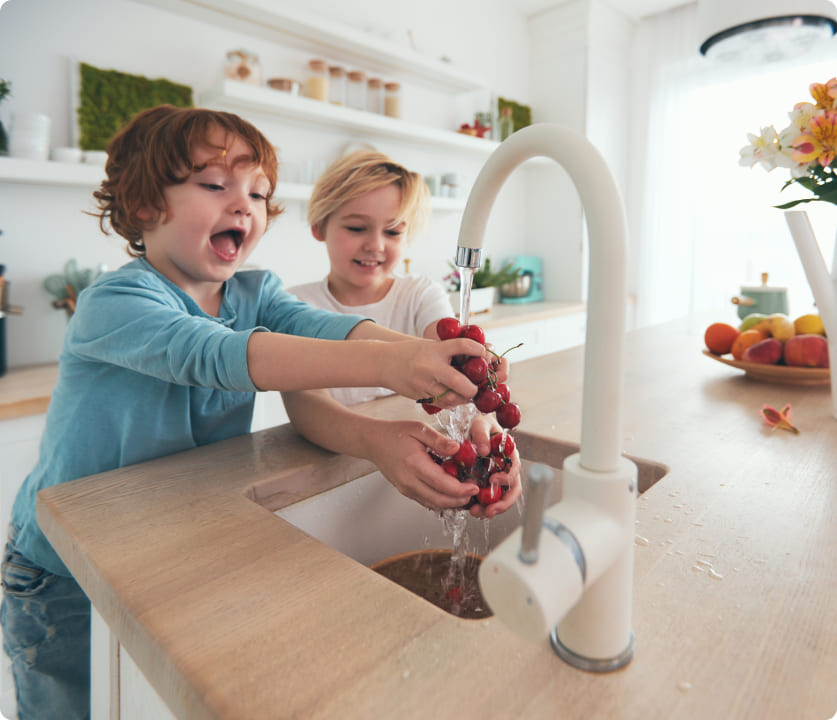 Two young children wash a bunch of red grapes under a kitchen faucet.