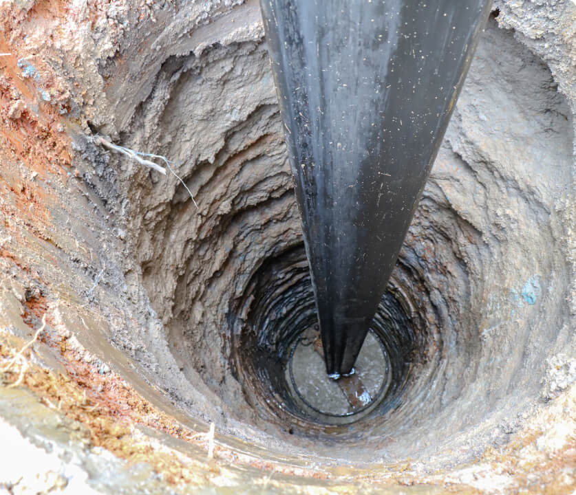Deep borehole with a vertical central pipe descending into layered soil during construction.