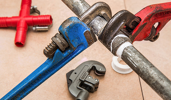Blue pipe clamped by a metal pipe wrench with red fittings and other plumbing tools on a workbench.