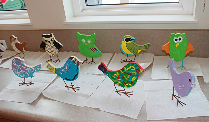 Colorful handmade paper birds with wire legs arranged on white sheets along a window.