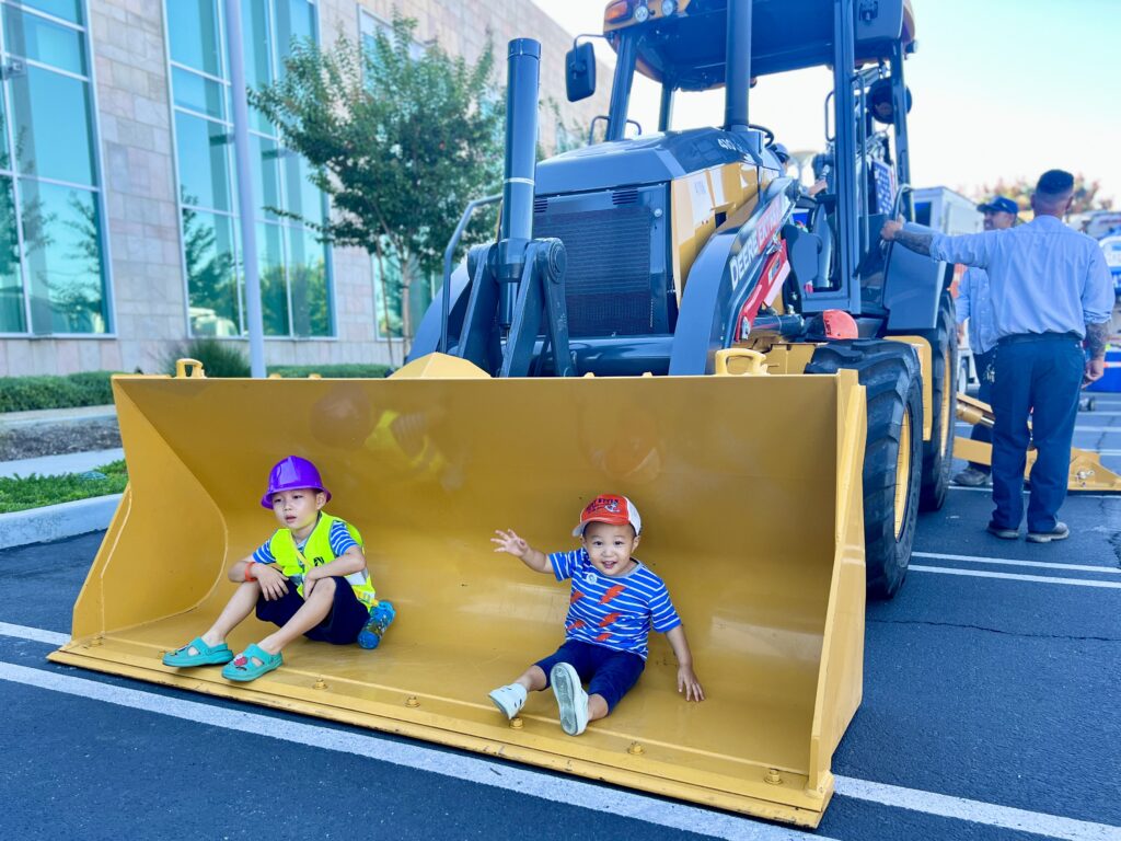 Two kids sit in the yellow front-loader bucket at an outdoor construction demo, wearing helmets beside a large tractor.