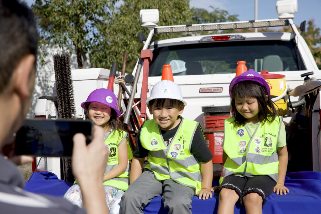 Three children in neon vests and hard hats sit on a blue platform in front of a white utility truck during a safety event.