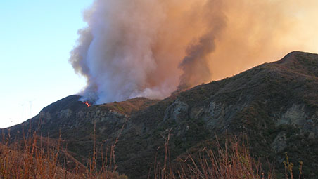 Smoke and orange flames rise from a hillside fire on a rocky ridgeline.