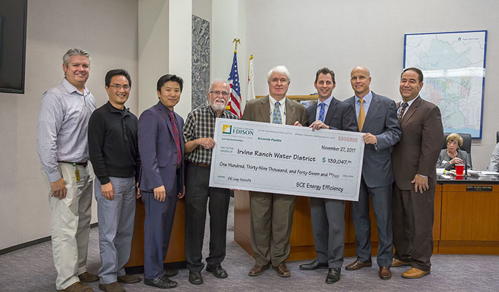Group of nine men in business attire standing in a conference room, holding a large ceremonial check.
