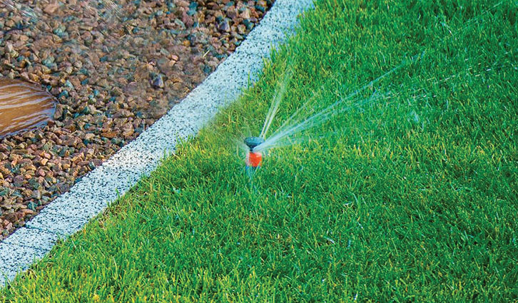 Garden sprinkler waters a green lawn beside a gravel border, with a visible water arc.