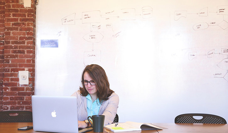 Woman with glasses working on a MacBook at a desk in a bright office with a whiteboard wall.
