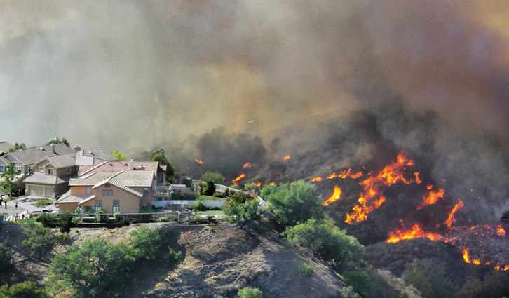 Residential hillside neighborhood with a wildfire burning nearby; flames and thick smoke fill the air.