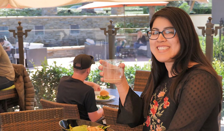 Smiling woman with glasses holding a glass at an outdoor cafe, with a plate of food on the table.