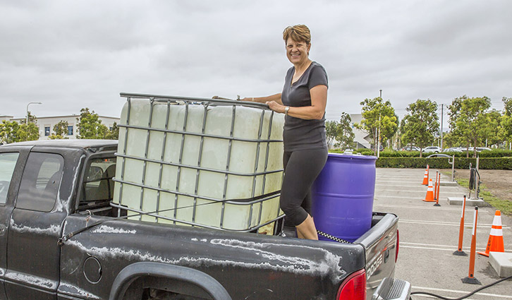 Woman standing in the back of a pickup truck loading a large IBC tote and a purple drum in a parking lot.