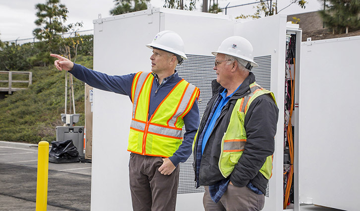 Two construction workers in hard hats and high-visibility vests discuss equipment outdoors, one pointing.