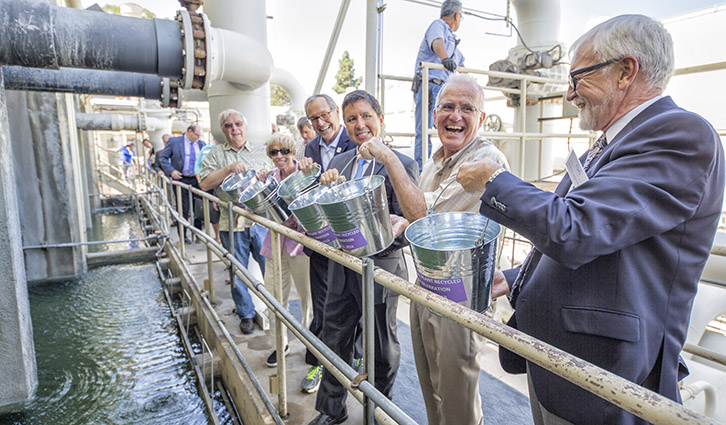 Group of adults on an industrial facility tour, holding metal buckets beside water and large pipes.