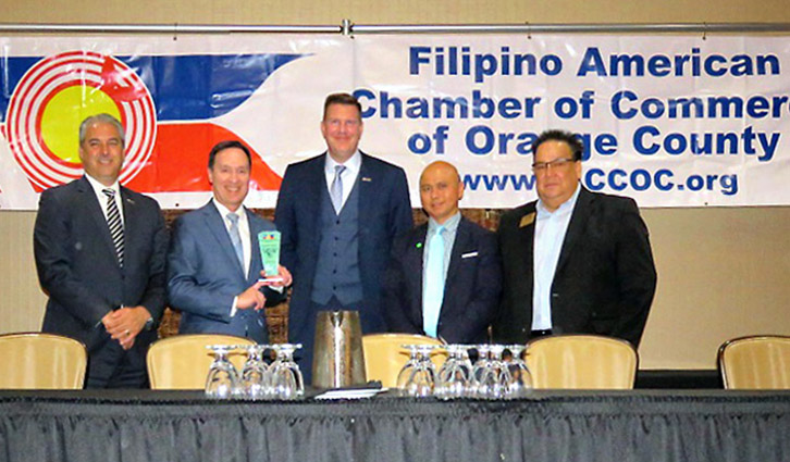 Group of five men in suits on a stage with a banner for the Filipino American Chamber of Commerce.