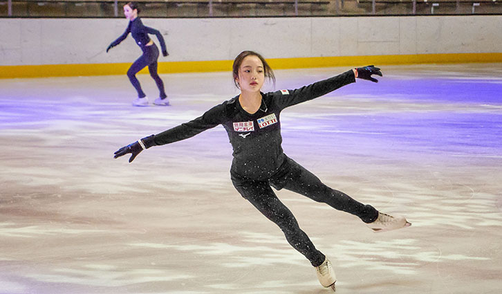 Female figure skater in a black costume performs a spiral on an ice rink; another skater glides in the background.