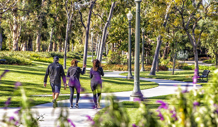 Three people walk along a curved park path among trees and lampposts, with flowers in the foreground.