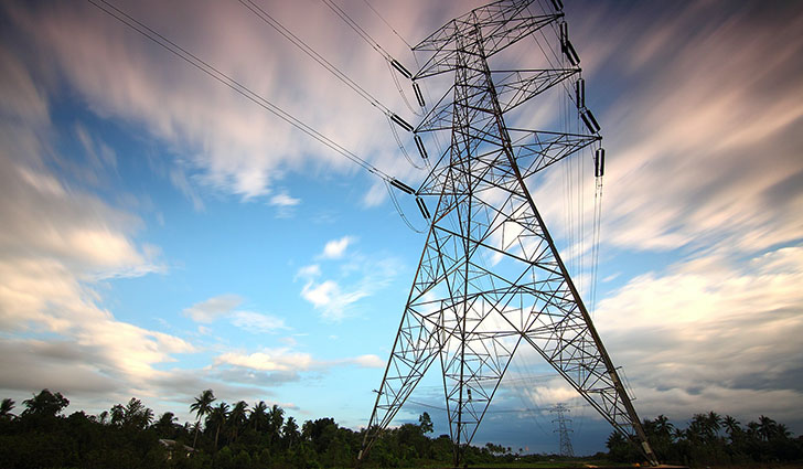 A tall steel lattice transmission tower with power lines stretches across a tropical landscape under a partly cloudy sky.