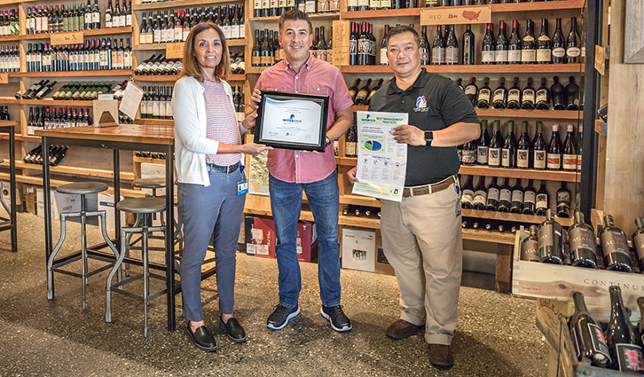 Three people in a wine shop pose with a framed certificate and a document.