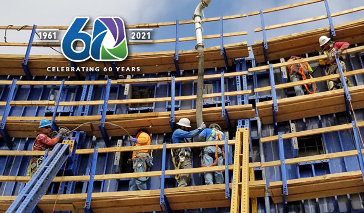 Construction workers in hard hats assemble blue wooden formwork on a building site, with a 60th anniversary logo in the upper left.