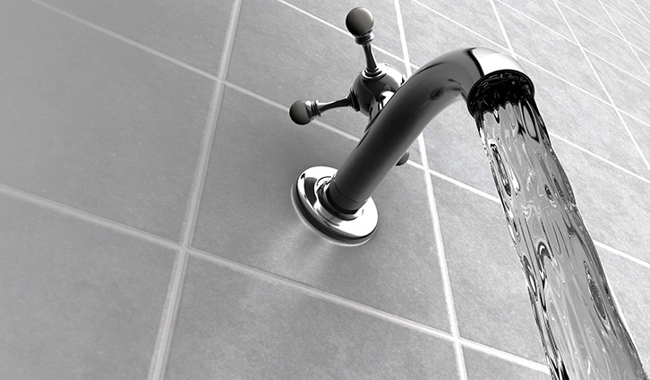 Chrome faucet with running water over gray tiled wall.