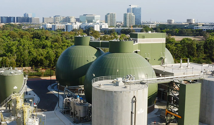 Green spherical anaerobic digesters at a wastewater treatment plant with a city skyline in the background.