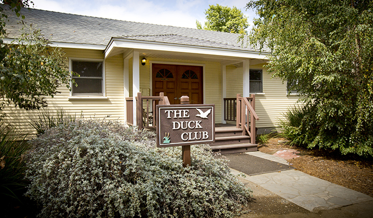 Front view of a beige single-story house with a wooden sign that reads The Duck Club in the front yard.